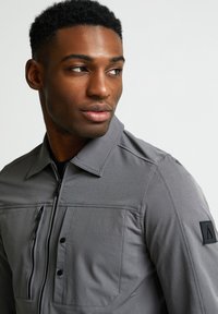 Pelēks jaciņa ar rāvējslēdzēju un augstu apkakli, ar divām krūšu kabatām, teksturētu audumu un melnu logotipa plāksni uz piedurknes.