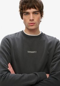 Mørkegrå sweatshirt lavet af bomuld, med en ribbet rund hals og et lille hvidt "SUPERDRY" logo på brystet. Glat tekstur.