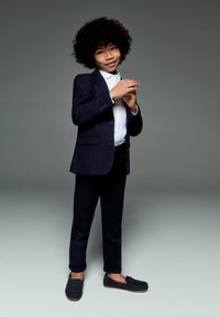 Un enfant portant un costume bleu marine foncé avec une chemise blanche, se tenant debout avec confiance. Il a des cheveux bouclés et des mocassins sombres, avec des pantalons retroussés pour ajouter une touche de détail.