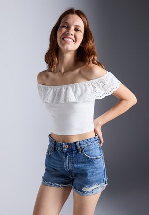 Top blanco sin mangas con volantes de encaje y un diseño corto, combinado con unos pantalones cortos de mezclilla azul desgastados. Detalles de tela texturizada visibles.