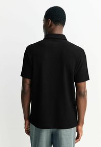 Sort ribbet kortærmet polo-shirt med klassisk krave, struktureret stof og lige kant, båret med lysegrå bukser.