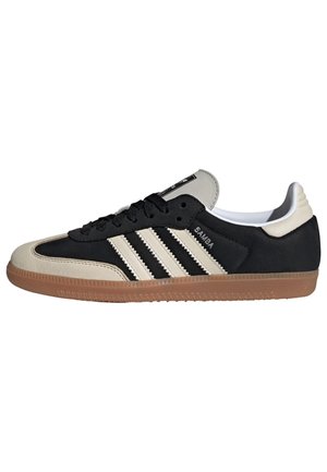 Baskets Adidas Samba noires et beiges avec semelle gomme, lacets noirs, trois bandes latérales beiges et texte "Samba" sur le côté.