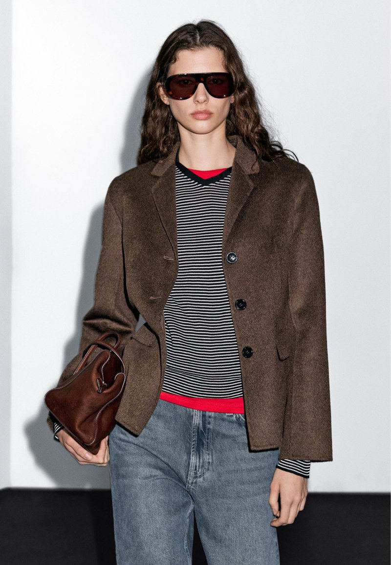 Blazer en laine marron avec poches latérales et deux boutons, superposé sur une chemise rayée noir et blanc. Accessoirisé avec un sac à main marron.