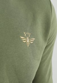 Pull vert olive avec une finition texturée et un logo d'abeille brodé en or sur le côté gauche. Design simple avec un col rond.