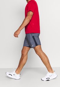 Man loopt in een rood T-shirt, grijze sportshorts met een zwarte streep en witte Nike sneakers tegen een effen witte achtergrond.