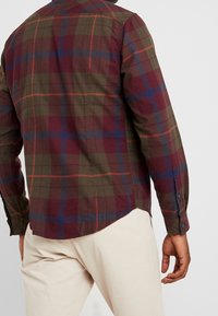 Camicia scozzese in verde scuro, borgogna e blu con maniche lunghe e polsini con bottone. Presenta un orlo curvo e una texture sottile.
