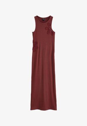 Donna in abito maxi senza maniche color ruggine che sta vicino a una finestra ad arco con vegetazione all'esterno, accanto a un divano bianco imbottito all'interno.
