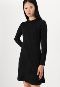 Vestido preto canelado com mangas longas, decote redondo, corte até ao joelho, apresentando um design moldado e um tecido suave e texturizado.