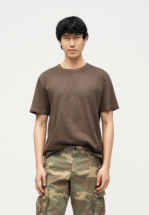 Fiatal fekete hajú férfi, barna pólót és terepmintás cargo nadrágot visel, egy egyszínű, világos háttér előtt áll.