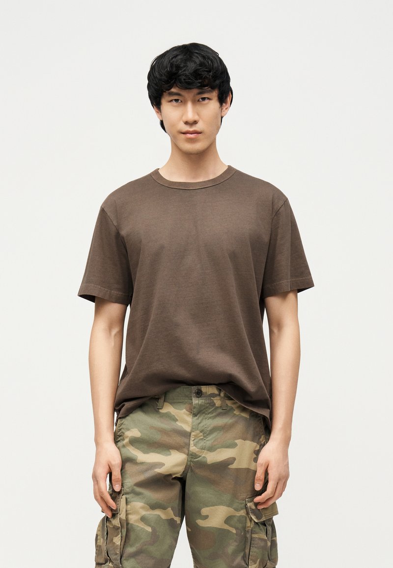 Νέος άνδρας με μαύρα μαλλιά, φοράει καφέ μπλουζάκι και καμουφλάζ παντελόνια τύπου cargo, στέκεται μπροστά σε απλό φωτεινό φόντο.