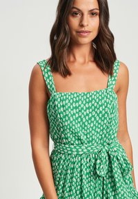Zelena cvetna obleka s kvadratnim izrezom, debelimi naramnicami in zategnjenim pasom. Narejena iz lahkotne tkanine, z gubanim teksturo in detajlom vrvice.