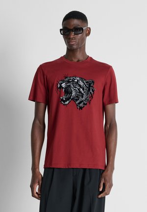Homme portant des lunettes de soleil foncées et un t-shirt rouge avec un motif léopard rugissant, associé à un pantalon noir, debout devant un fond uni.