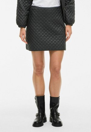 Femme portant une mini-jupe matelassée noire, des manches de veste matelassée assorties et des bottes en cuir noires jusqu'au mollet avec des boucles.