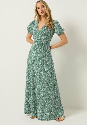 Vrouw draagt groene bloemenprint wikkel maxi jurk met pofmouwen en strikceintuur, staat met handen op de heupen.