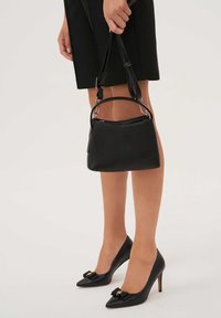 Črna usnjena torbica z zaobljeno obliko, z dvema ročajema, z zapiranjem z zadrgo in subtilnim odtisnjenim logotipom. Črne čevlje z visokimi petami in poudarkom v obliki pentlje.