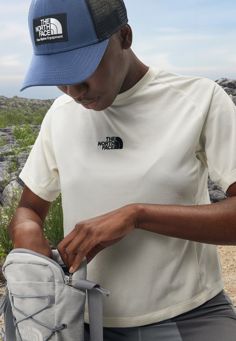 Košulja kratkih rukava svijetlo bež boje s crnim logotipom "The North Face", u kombinaciji s sivom torbicom i plavo/crnom mrežastom kapom, na otvorenom s stijenama u pozadini.
