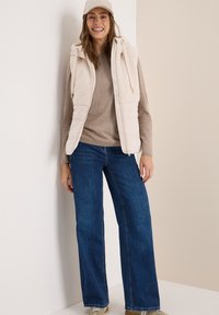 Beige ärmlös väst med en texturerad, fluffig ovandel, bärs över en ljusbrun tröja, tillsammans med vida blå jeans och en beige keps.