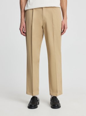 Mężczyzna ubrany w beżowe spodnie o prostych nogawkach, białe skarpetki i czarne skórzane mokasyny, stojący na tle jednolitego jasnego tła.