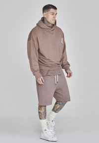 Sudadera con capucha marrón y logo frontal, combinada con pantalones cortos a juego. El modelo lleva zapatillas blancas y calcetines, mostrando tatuajes en los brazos y las piernas.