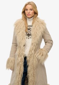 Beige yderjakke med en lang, shaggy faux pelskrave og manchetter. Den indre sweater har et strikket mønster i jordnære farver.