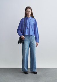 Mujer de pie contra un fondo liso, vistiendo una camisa de rayas azul con botones, jeans anchos azules con pequeños adornos, zapatos negros y un bolso de hombro negro.