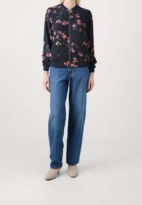 Giacca bomber con zip floreale in blu navy con fiori rosa, abbinata a jeans a vita alta, morbidi e chiari e stivali alla caviglia beige.