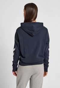 Kvinna med långt brunt hår som bär en marinblå huvtröja med huvan nedfälld och ljusgrå mjukisbyxor, sedd bakifrån mot en enfärgad bakgrund.