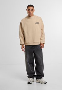 Felpa in cotone beige con collo alto, vestibilità oversize e stampa sul lato sinistro del petto. Abbinata a pantaloni in denim neri larghi e scarpe da ginnastica.