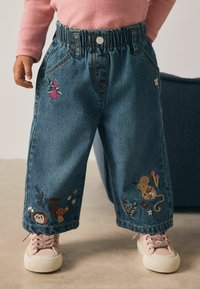 Kind trägt eine weite Jeans mit bestickten Blumen, Pilzen und einer Maus, kombiniert mit rosa Turnschuhen und einem rosa Pullover.