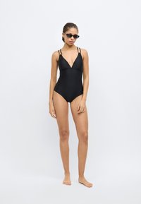 Juodas vientisas maudymosi kostiumėlis su gilia iškirpte, reguliuojamomis petnešėlėmis ir elegantišku dizainu. Dėvimas su mažomis juodomis saulės akiniais.