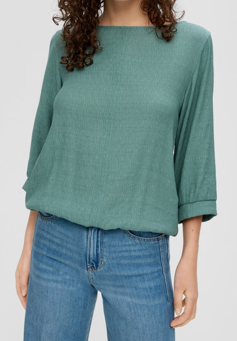Femme portant un chemisier vert à manches longues et col bateau associé à un jean denim bleu clair sur un fond uni.