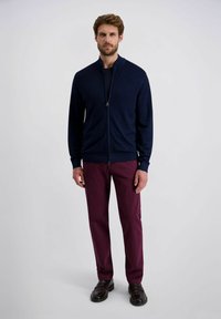 Marineblauer Reißverschluss-Pullover mit Stehkragen, strukturierter Stoff. Getragen mit bordeauxfarbenen, schmal geschnittenen Hosen und dunklen Schuhen, stehend vor einem neutralen Hintergrund.