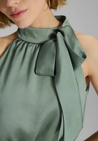 Groene blouse met textuur, hoge halslijn en een grote strikaccent op de schouder, met geplooide details en zachte stof.