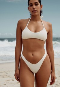 Biely dvojdielny bikini vyrobený z textúrovanej látky. Top s halter krkom a trojuholníkovými košíčkami, spodný diel v štýle tang s nízkym pásom a výstrihom do tvaru V.