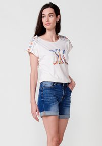 T-shirt beige clair à manches courtes avec broderies colorées et accents de manches détaillés, assorti à un short en denim bleu à ourlet retroussé.