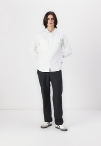 Chemise blanche à boutons avec deux poches frontales, associée à un pantalon gris foncé et des baskets blanches avec des accents noirs. Fond uni.