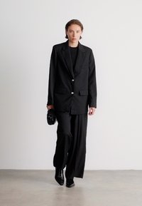 Sacou negru, croit, cu două butoane, detalii pe buzunare, asortat cu pantaloni negri largi. Modelul poartă o mică poșetă neagră și pantofi cu vârf ascuțit.