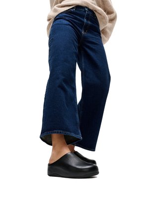 Persoon die donkerblauwe jeans met wijde pijpen en zwarte instapschoenen draagt, staand tegen een witte achtergrond.