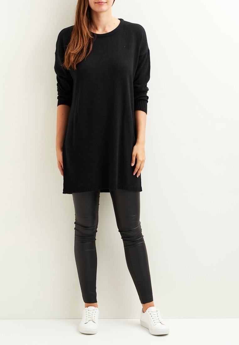 Femme debout portant un pull tunique noir à manches longues, des leggings en cuir noir et des baskets blanches sur un fond clair uni.