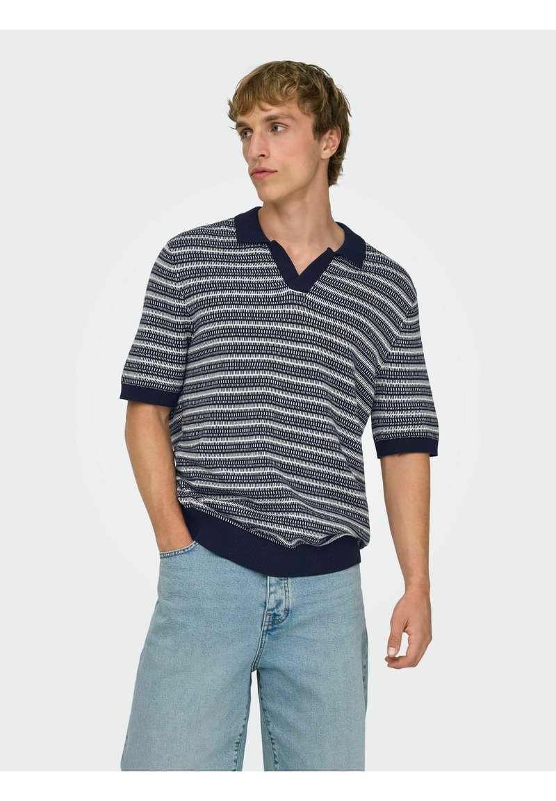 Homme portant un polo à manches courtes rayé bleu marine et blanc avec col en V et jean bleu clair, debout avec une main dans la poche.