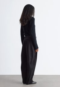 Schwarzer, flauschiger Pullover, hoch geschnittene schwarze Hose mit weitem Bein, glatte Textur, minimalistische Details und eng anliegende schwarze Schuhe.