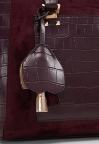 Bordeaux håndtaske med en kombination af glat og struktureret læder, med en blank krokodillepræget detalje og en metallisk gylden frynse-accent.