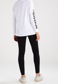 Biała bluzka z długim rękawem z napisem "Converse" wydrukowanym pionowo na czole w czerni, zestawiona z czarnymi legginsami i jasnymi sneakersami.