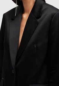 Černý blazer s klopou, jedním knoflíkem a strukturovaným střihem. Hladká textura s minimalistickým designem a jemnými švy.