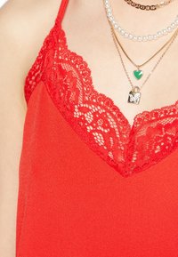 Top sin mangas de color rojo con un escote en V de encaje. La tela es suave y presenta finos detalles de encaje en los bordes. Tres collares en capas decoran el escote.