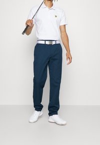 Vit polo-shirt med en gul logga, marinblå byxor, vit bälte och vita golfskor. Golfklubba hålls i ena handen.