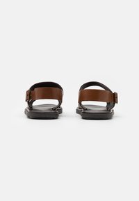Braune Ledersandalen mit schwarzer Sohle, ausgestattet mit zwei Riemen und einem Schnallenverschluss. Minimalistisches Design mit flacher Sohle und offenen Zehen.