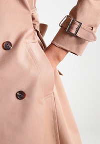 Beżowy płaszcz trench jest zapinany na podwójne guziki, posiada pasek z metalową klamrą oraz boczną kieszeń. Materiał ma gładką, matową fakturę.