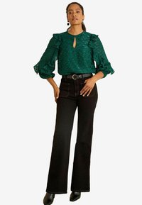Blusa verde con trama, maniche a balze e dettaglio a chiave, abbinata a jeans neri a zampa d'elefante e scarpe nere, accessoriata con una cintura.