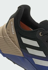 Chaussure de sport noire avec tige en mesh, accents beige et bleu, dotée de trois rayures blanches et d'une semelle texturée pour la traction.